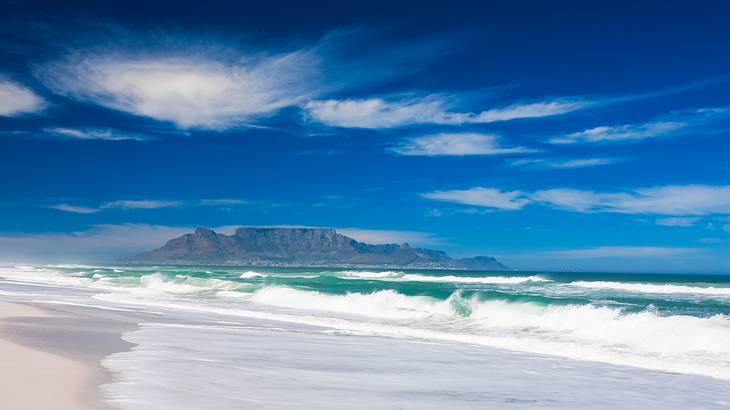 Waves crashing on a white beach with a view of a flat-topped mountain in the back