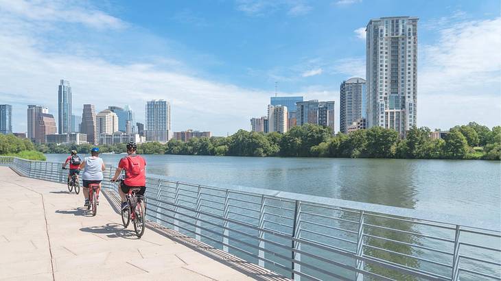 Three people biking on a path next to a lake and highrise buildings