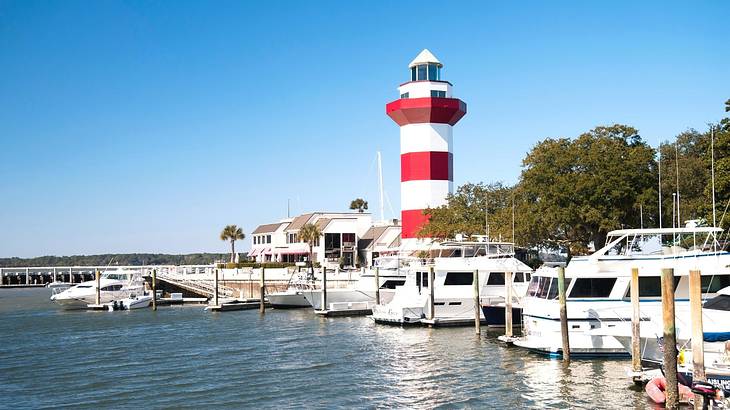 A marina with boats docked next to a white and red striped lighthouse and trees