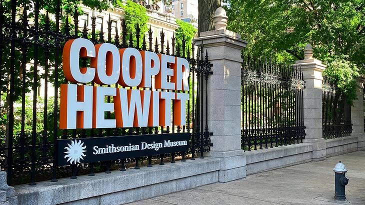 An iron gate and stone pillars with a white and orange sign that says "Cooper Hewitt"