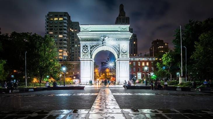 A stone arch with trees and buildings behind it and a road in front at night