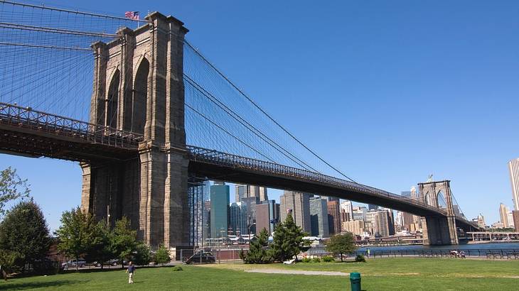 A suspension bridge with a park at the bottom and a river and skyline behind it