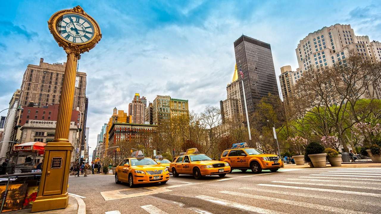 A street with trees and buildings, yellow taxi cabs on the road, and a gold clock