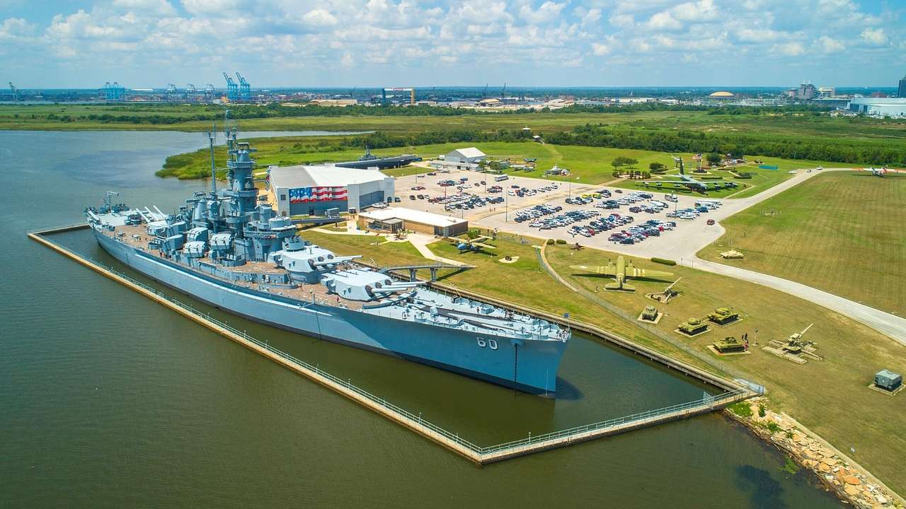 A baby battleship in a bay with grass and a building and parking lot to the side