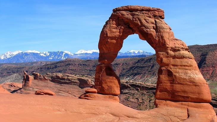 A red rock arch on a red rock cliff and snow-capped mountains in the distance