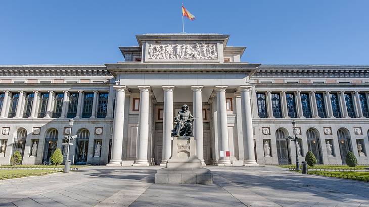 A bronze statue on a pedestal in front of a building with 6 columns and a flag on top