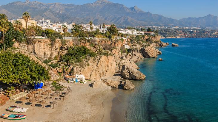 Boats and beach umbrellas on sand, houses on a cliff, and mountains at the back