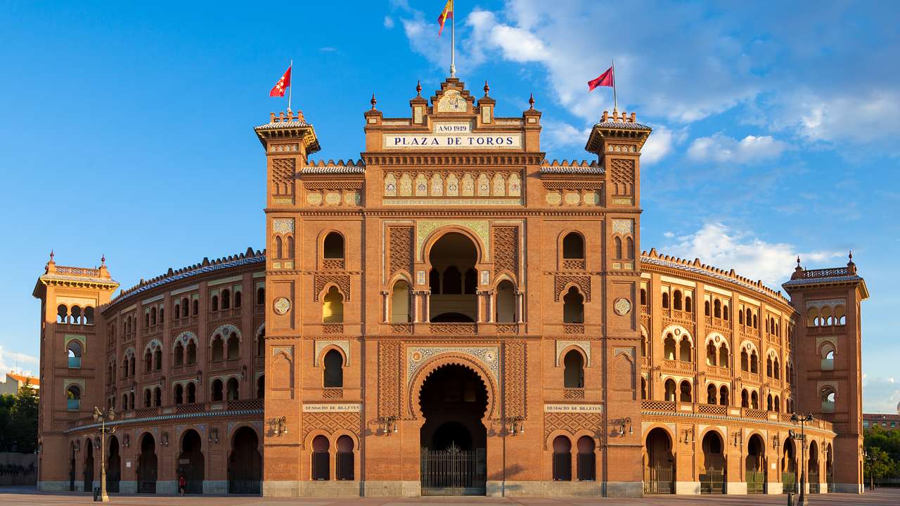 The facade of a bullring with three flags on top against a partly cloudy sky