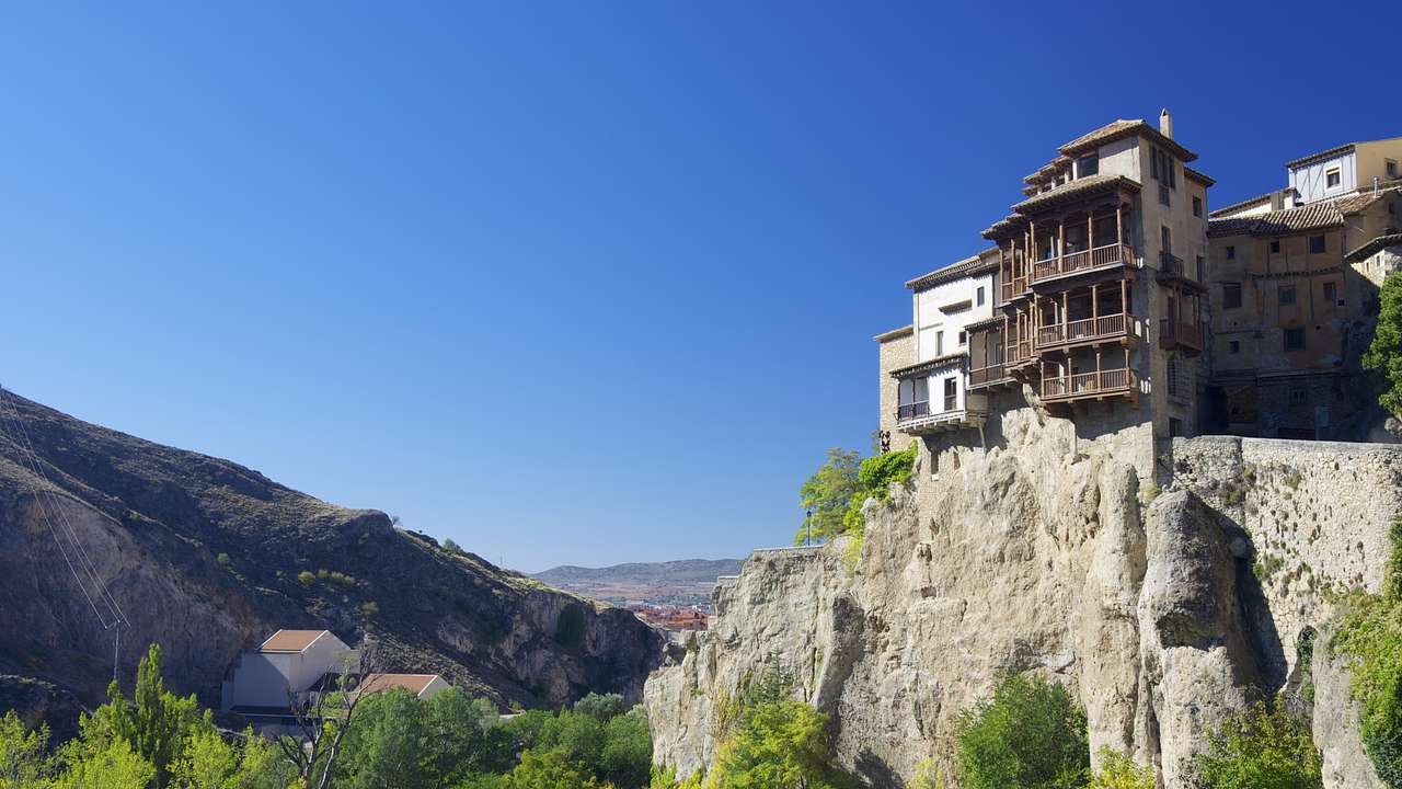 Houses built on a rocky cliff, trees below, and mountains at the back on a clear day