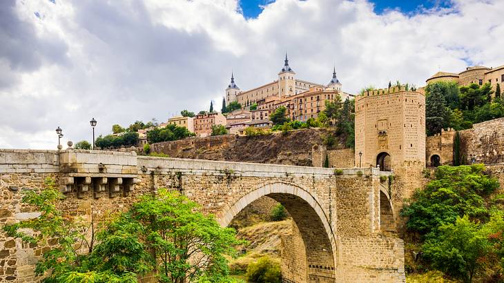 A bridge in the foreground of a fortress sitting atop a hill surrounded by greenery