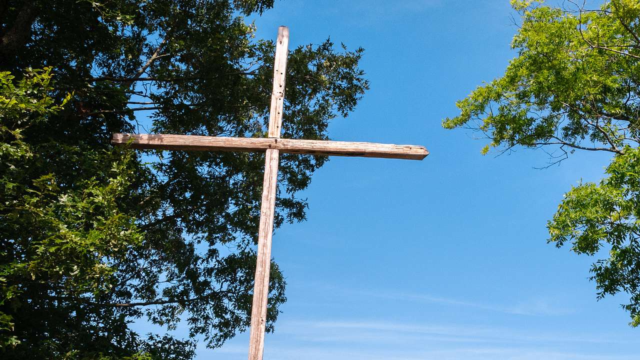 View from below of a wooden cross surrounded by greenery and a lake in the background
