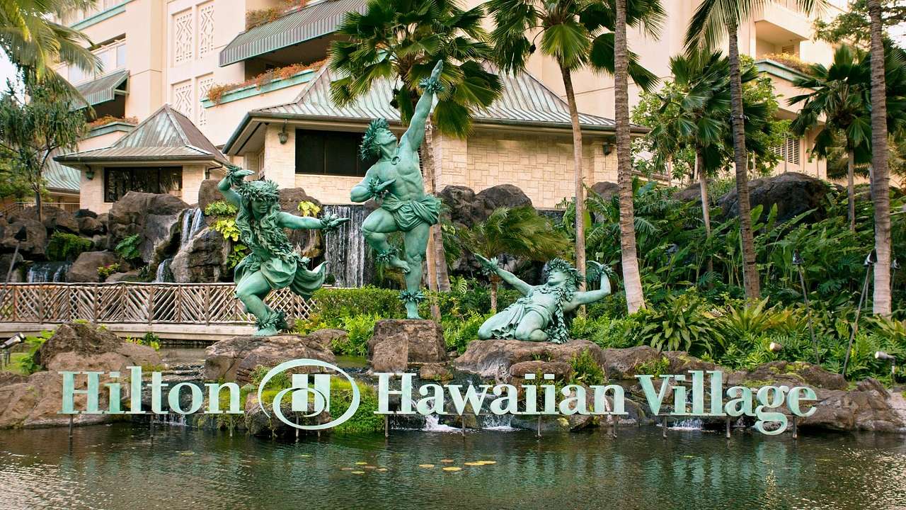 A sign that says "Hilton Hawaiian Village" with green statues behind it