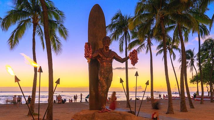 A statue of a man next to a surfboard with palm trees around it at sunset