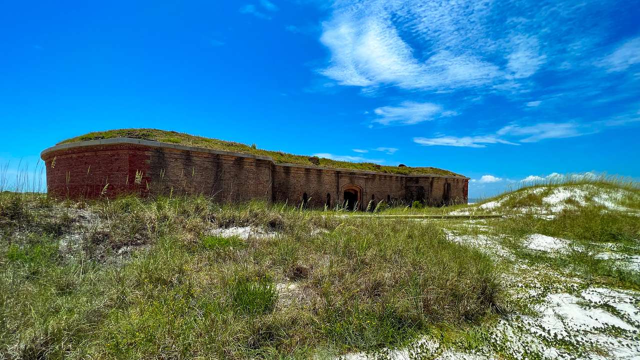 A one-story brick fort on a sandy shore filled with plants on a partly cloudy day