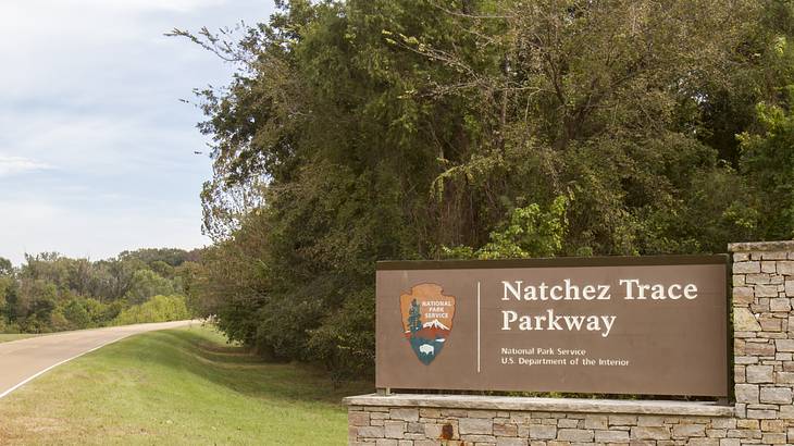 Brown signage on top of layered bricks on a grassy field with trees at the back Brown signage on top of layered bricks on a grassy field with trees at the back
