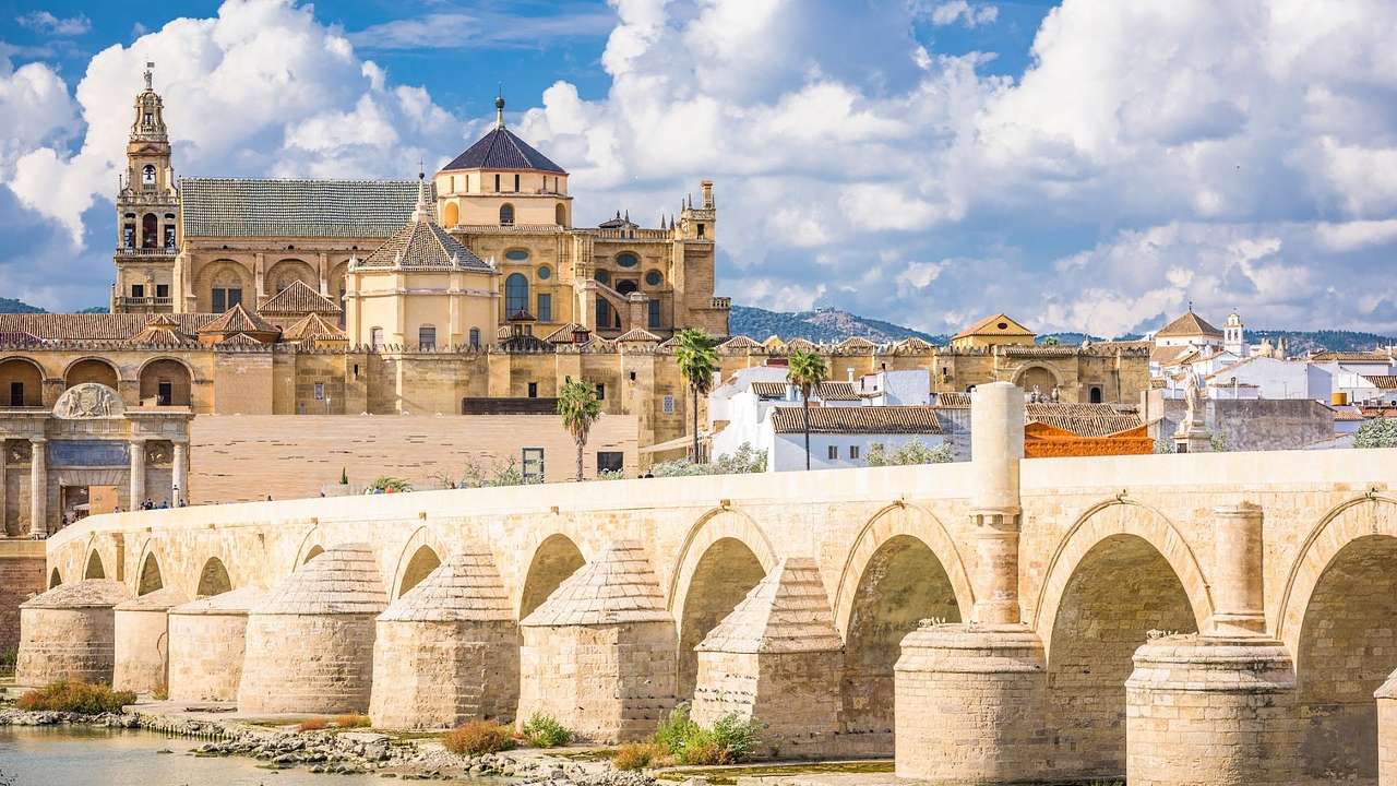 A stone bridge with many arches over a river next to ancient Spanish buildings