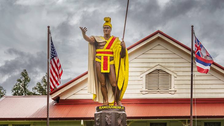 A bronze statue with a yellow cloak between the flags of the US and Hawaii