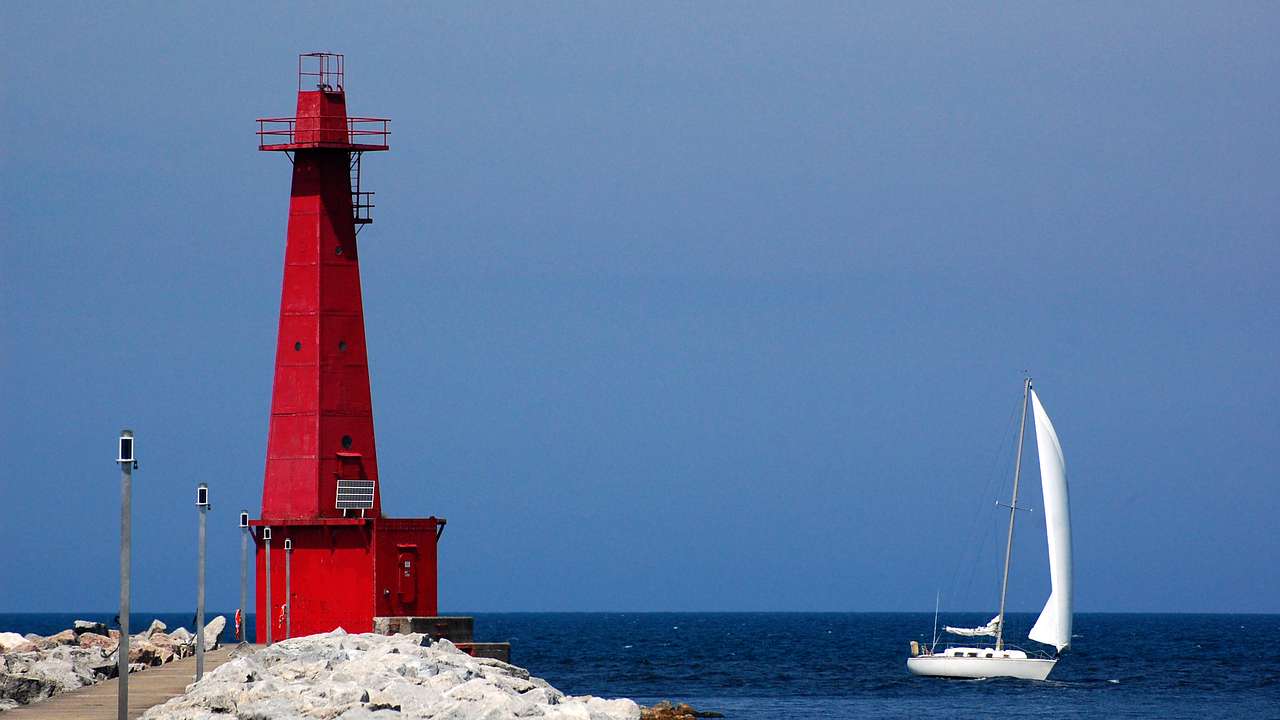 A white sailboat on a lake and a red lighthouse on its rocky coast