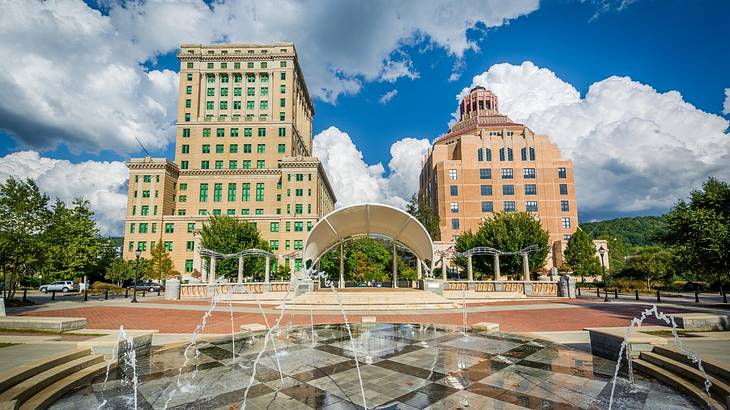 A city square with a fountain next to two buildings under a blue sky with clouds