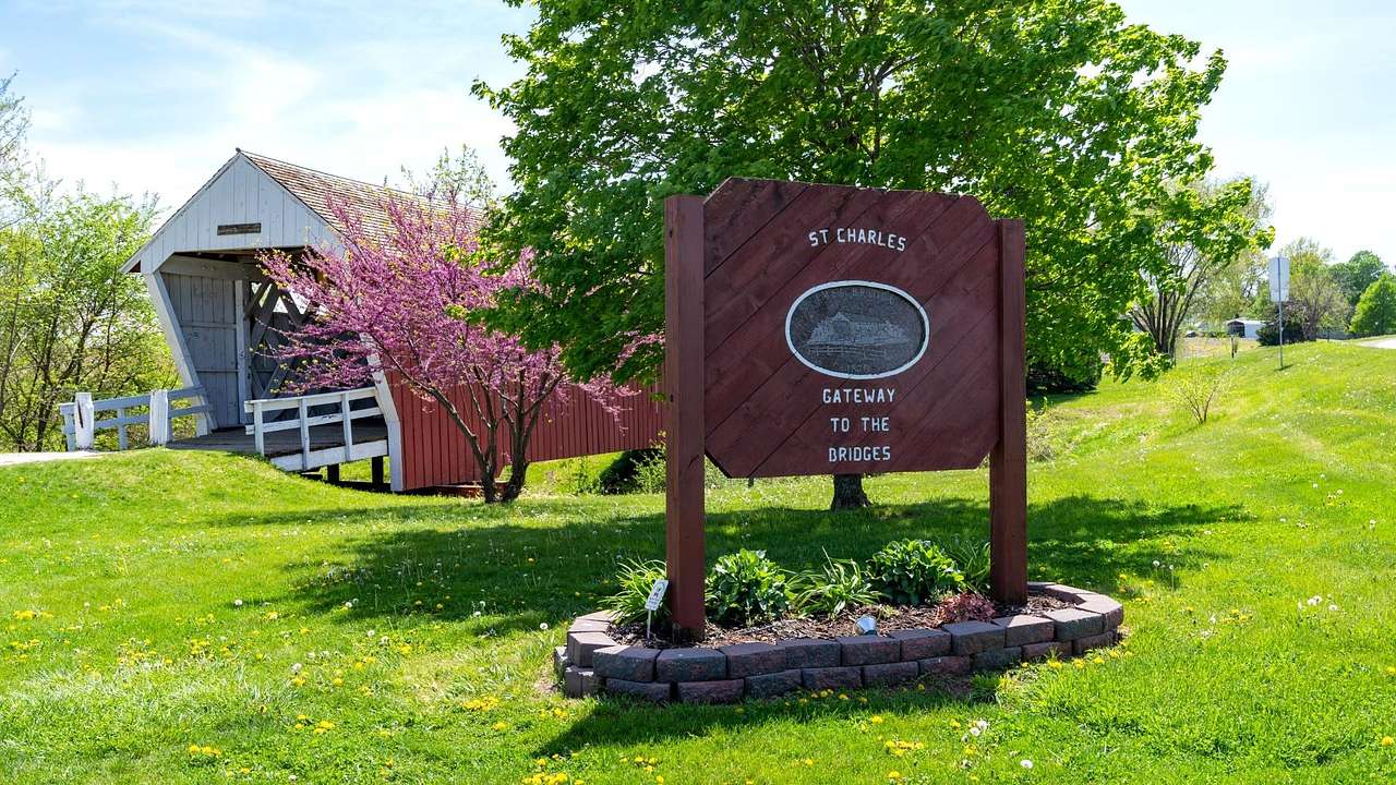 A red sign on green grass against a red covered bridge