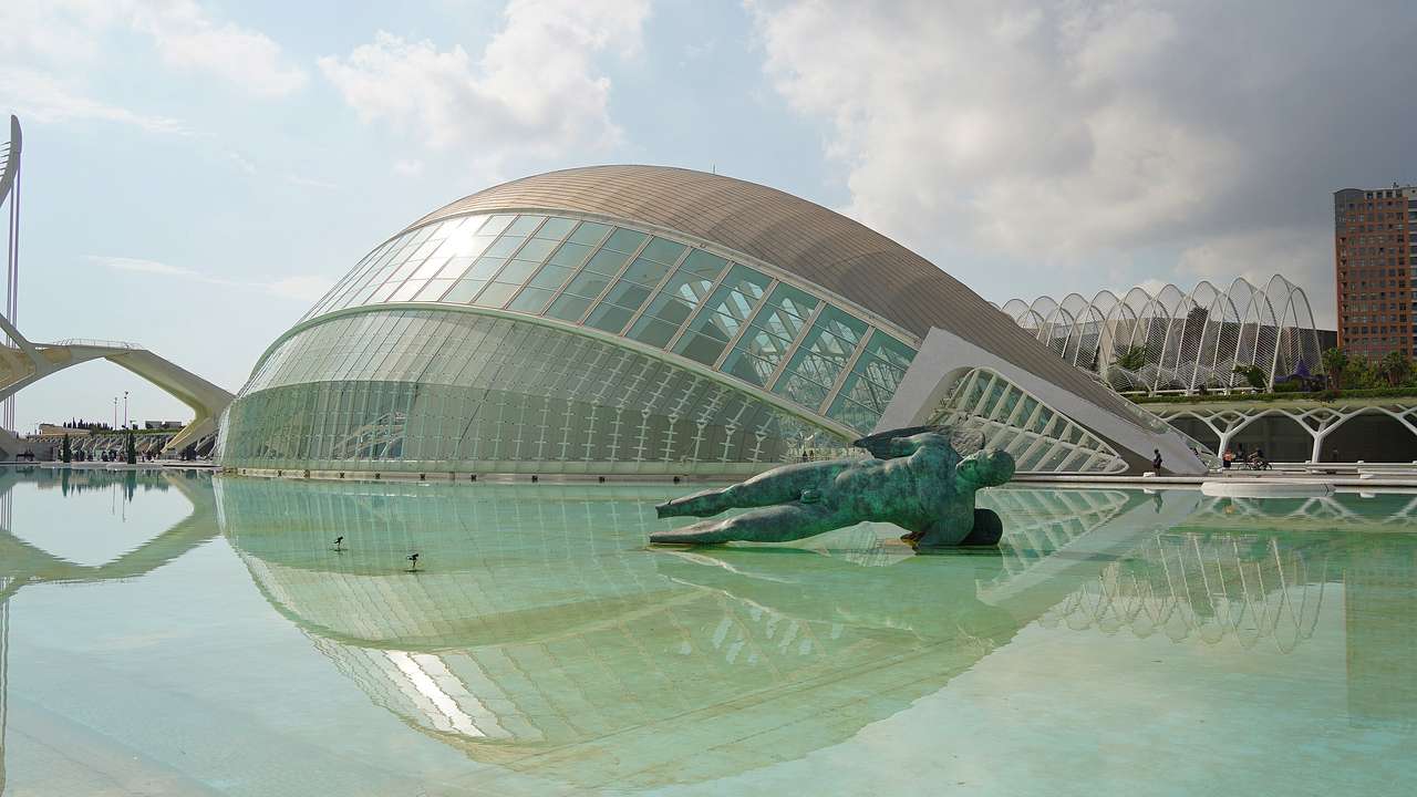 A modern half-dome building behind a fallen Roman man statue in shallow water