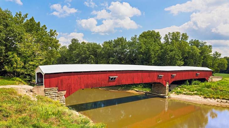A red covered bridge with a white roof over water against green trees