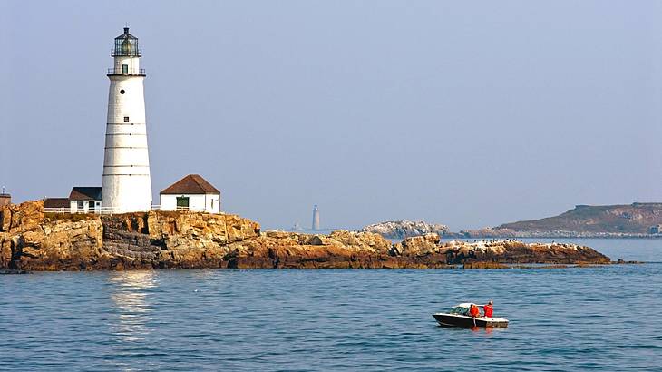 A white lighthouse on a rocky shore overlooking the water with a white boat