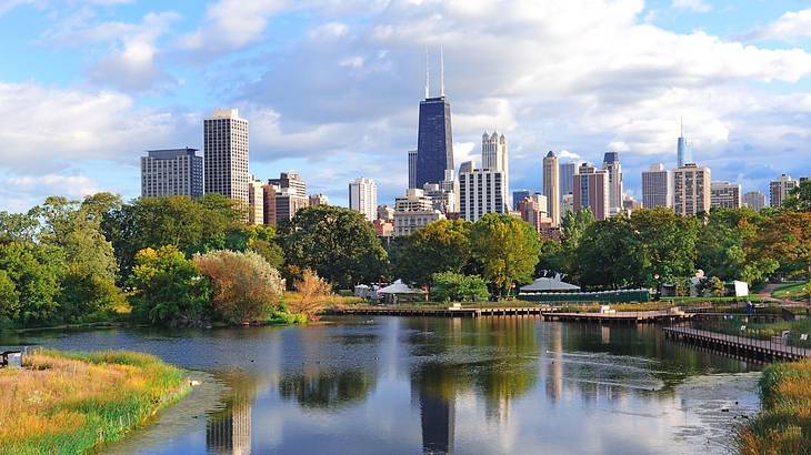 A pond surrounded by greenery in front of a city skyline on a slightly cloudy day