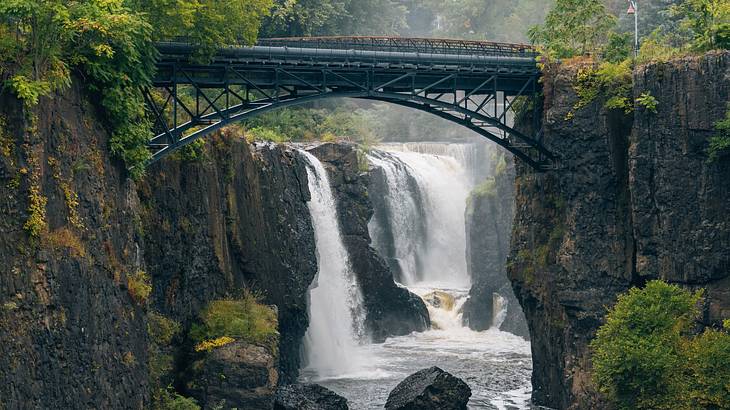A steel footbridge and a waterfall emptying into a river surrounded by greenery