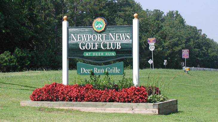 A sign on the grass that says "Newport News Golf Club" with red flowers below it