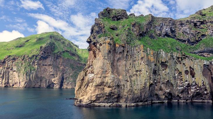 Rocky cliffs with greenery on them surrounded by water under a blue sky with clouds
