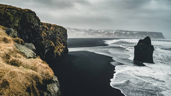 A black sand beach next to the ocean and rocky cliffs under a grey, cloudy sky