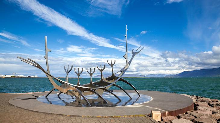 A metal ship-like sculpture next to the water under a blue sky with clouds