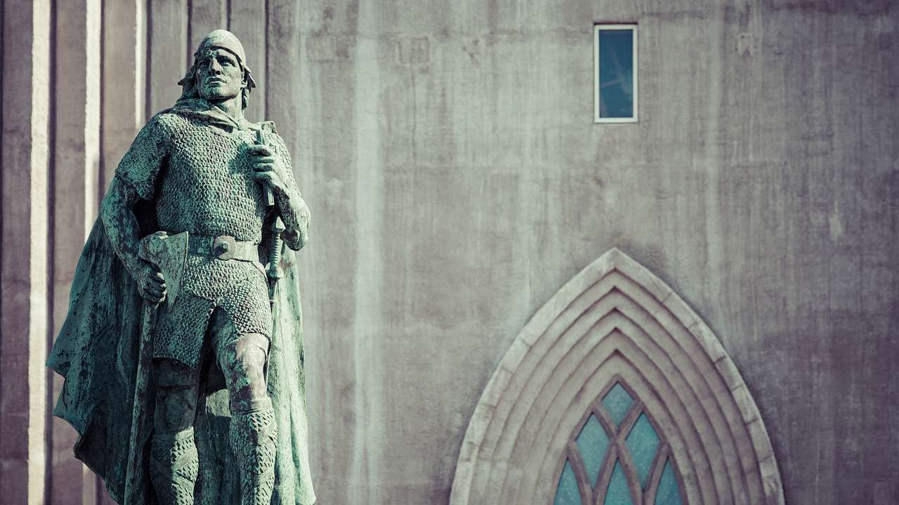 An oxidized statue of a man in armor with a sword in front of a stone building