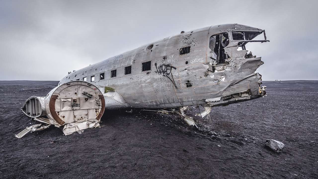 The wreckage of an aircraft on a black beach under a grey, cloudy sky
