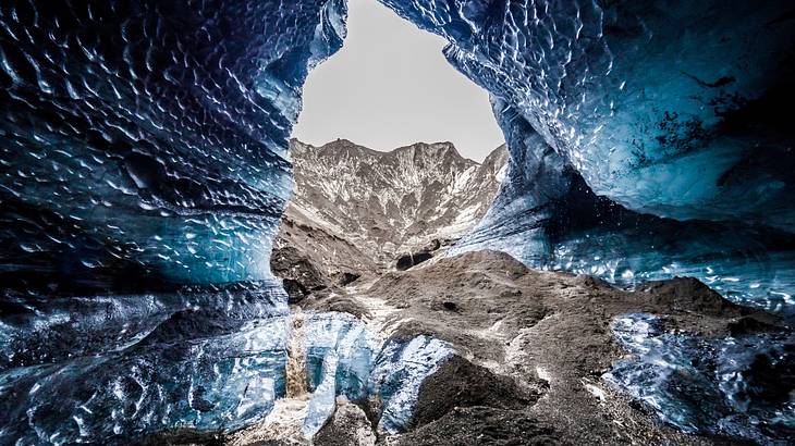 The interior of a purple-black glacial cave with a lookout to the sky and a mountain