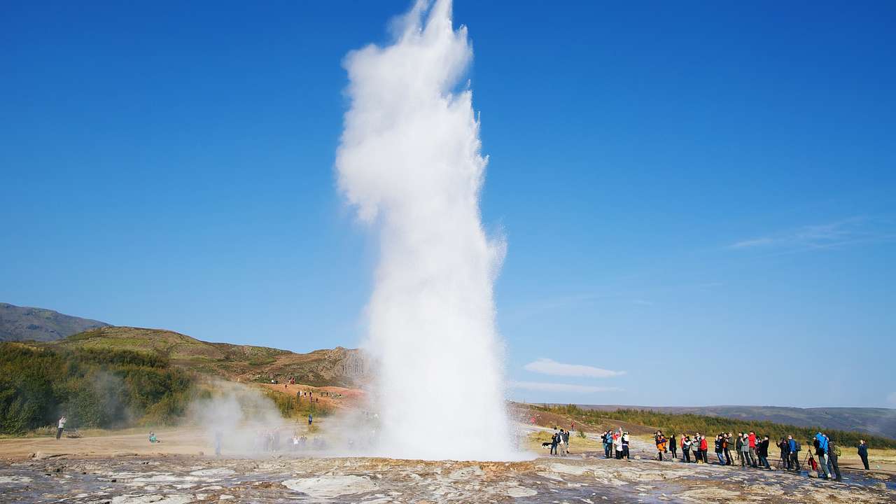 A geyser erupting with hills around it under a clear blue sky