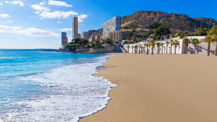 A sandy beach with gentle waves on the shore next to buildings and a hill