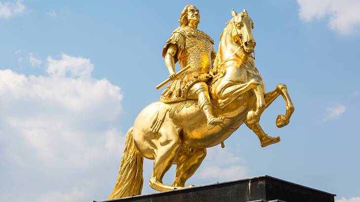 A golden statue of a man in armor on a horse in front of a blue sky with clouds