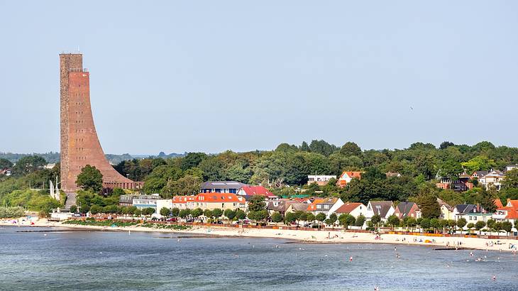 A curved red brick structure next to trees, small houses, a beach, and the water