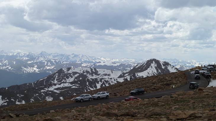 The summit of a mountain with cars on a road and snow-capped mountain ranges behind