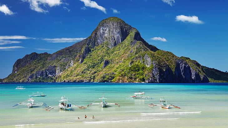 A greenery-covered hill next to turquoise water and a white sand beach with boats