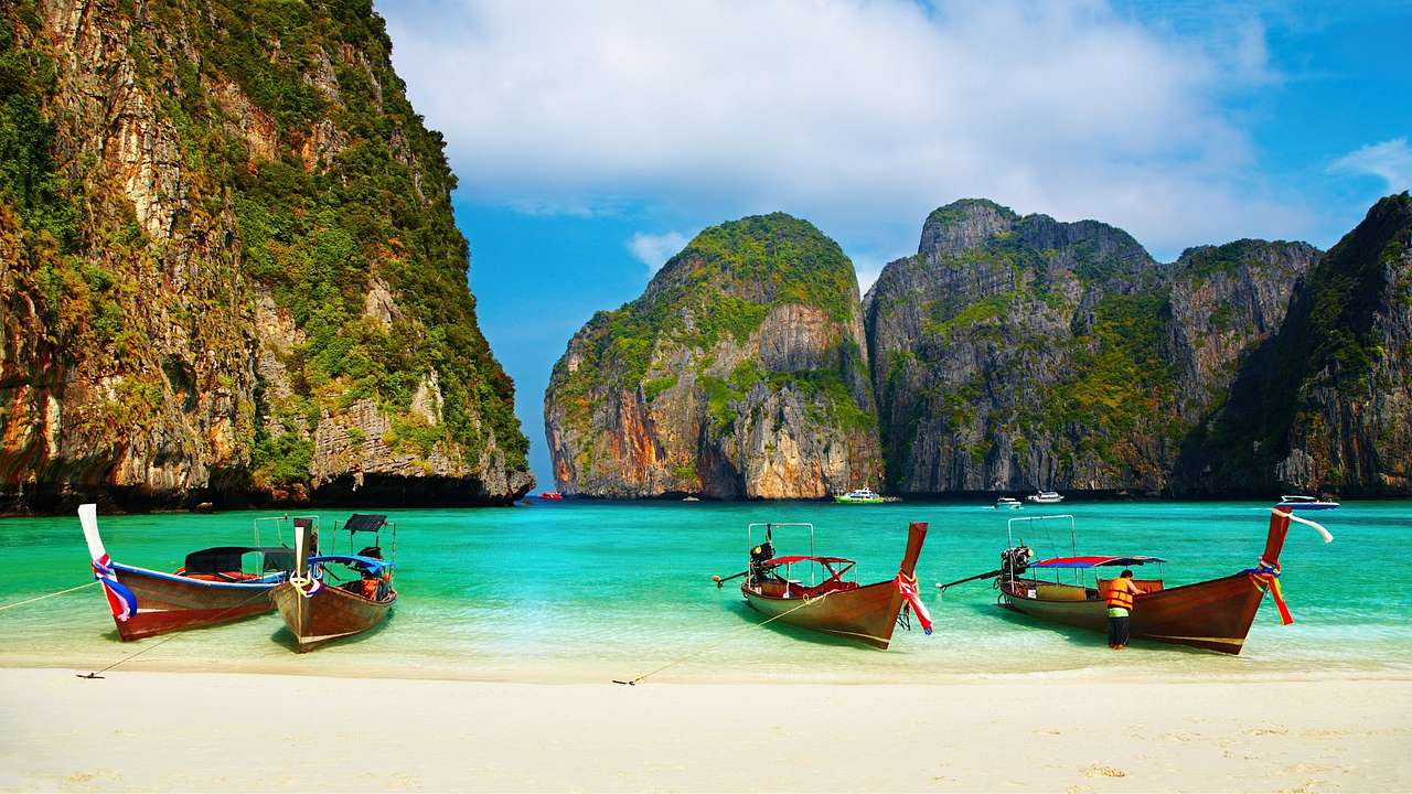 Thai wooden longtail boats docked on a white sand beach next to rocky green cliffs