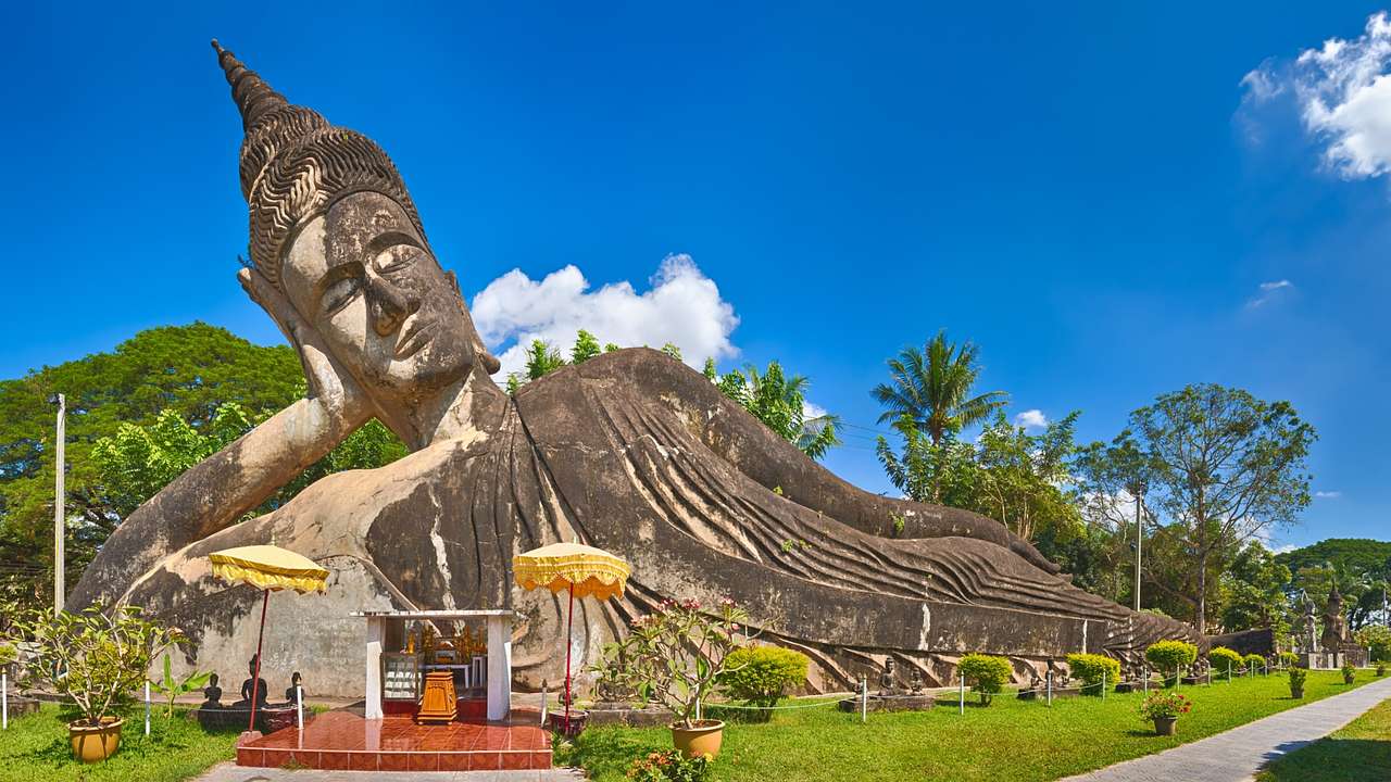A statue of a reclining Buddha surrounded by greenery under a blue sky