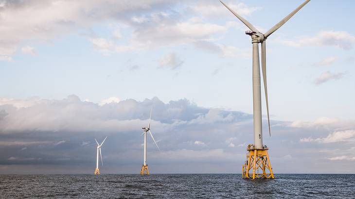 Three large wind turbines rising above the sea against a cloudy, light blue sky