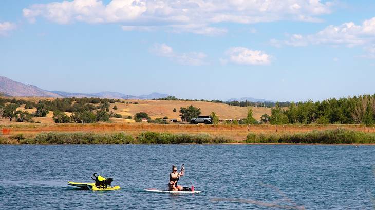 A person on a paddleboard on a lake next to hills and trees under a blue sky