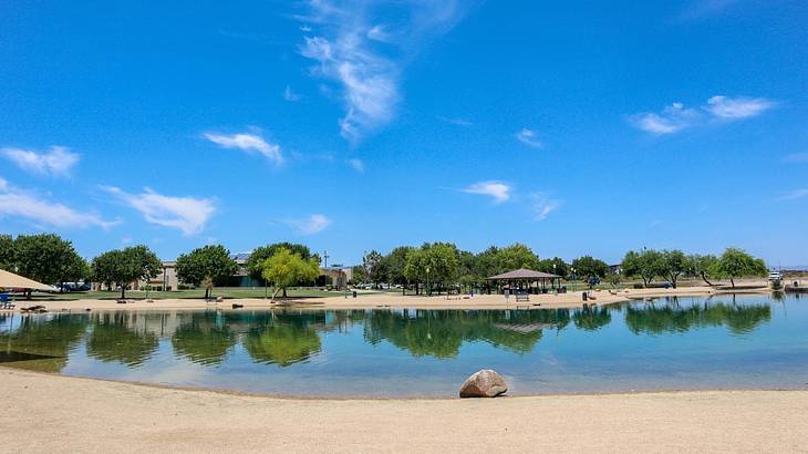 A pond next to a sandy shore with green trees on one side under a blue sky