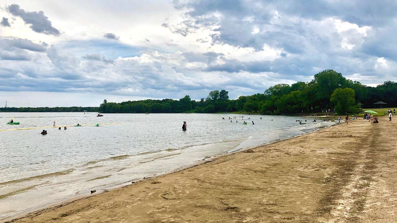 A lake next to a sandy shore and green trees under a cloudy sky