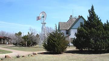 An American windmill next to a small house, grass, and trees under a blue sky