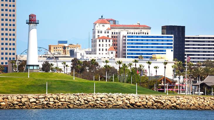 Water next to a stone wall, grass, a lighthouse, and buildings on a clear day